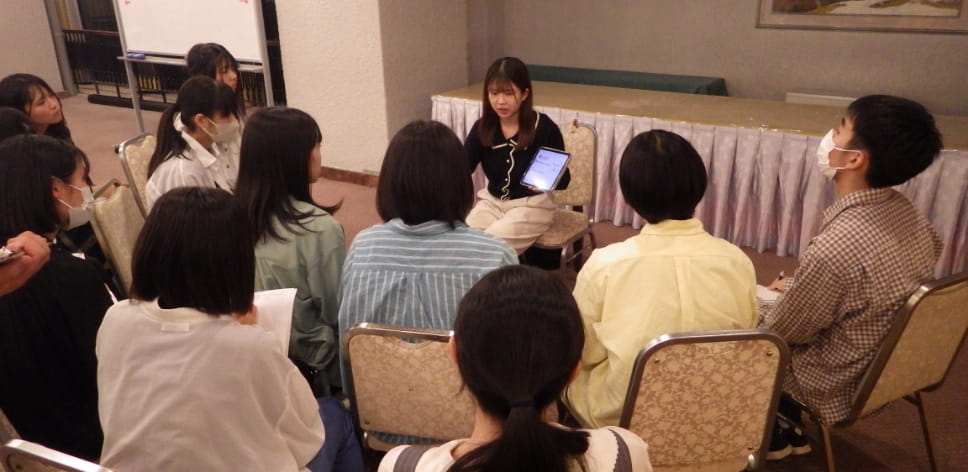 円形に座った学生たちが、タブレットを持った発表者の話を熱心に聞いている様子