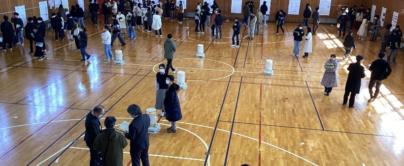 複数のストーブが配置された体育館内で多くの人が集まり、展示されたポスターを見ながら議論している様子