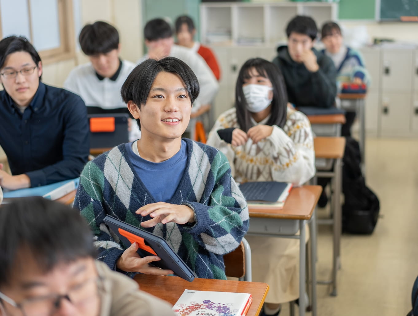 教室でタブレットを持ちながら発言する学生と、それを聞くクラスメートたち。