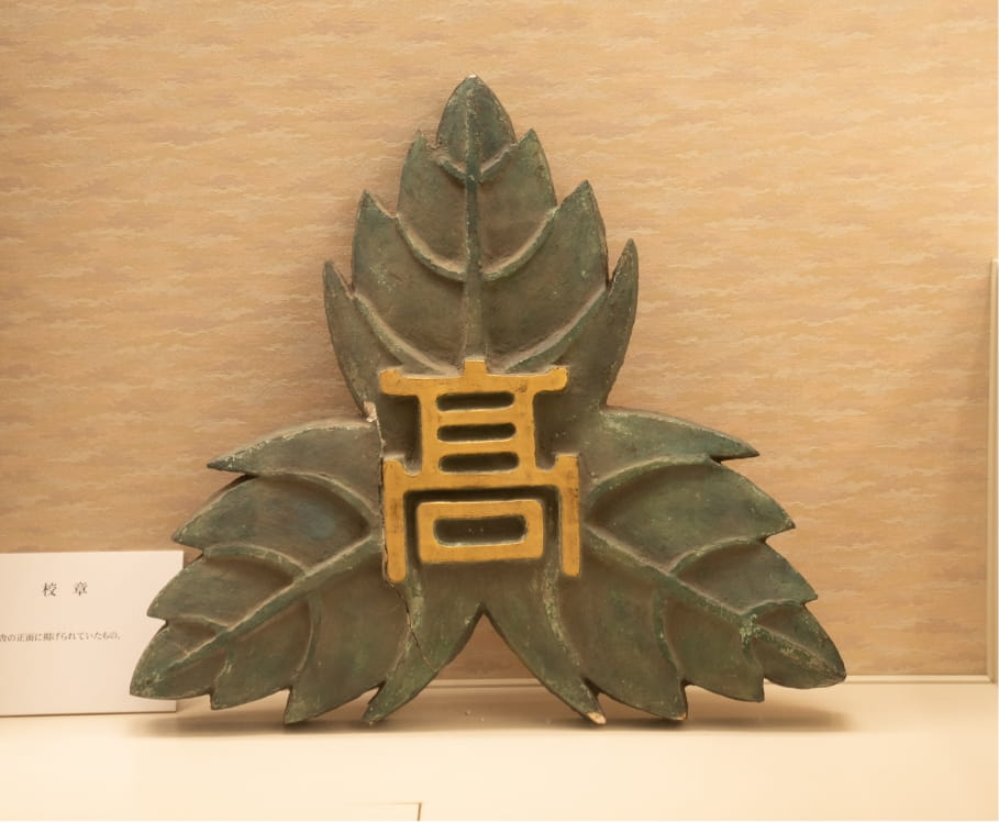 葉の形をした金属製の長岡高校の校章で、中央に「高」の文字がデザインされている。
